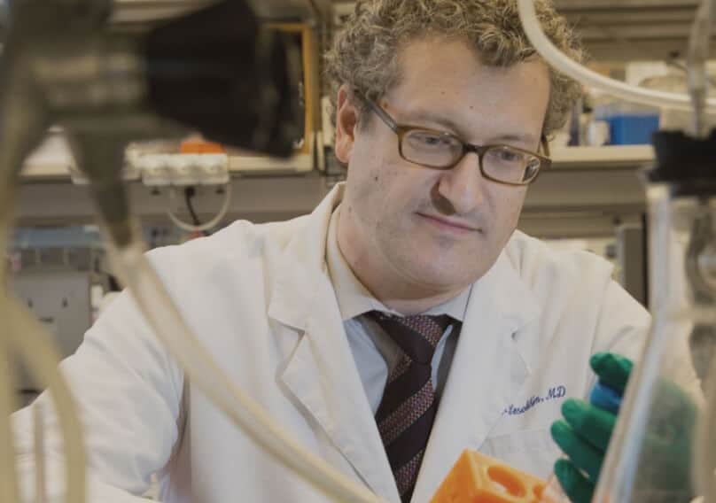 A scientist, surrounded by lab equipment, holds a vial.