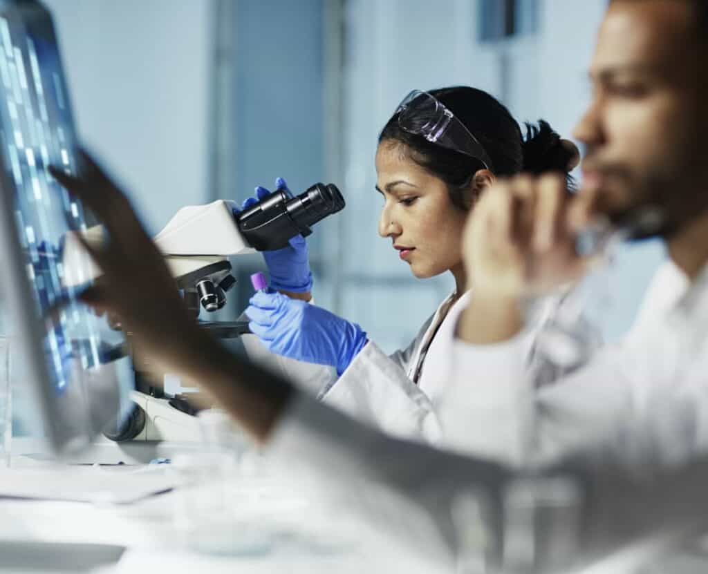 Two people in a lab, one studying a microscope, the other looking a computer screen